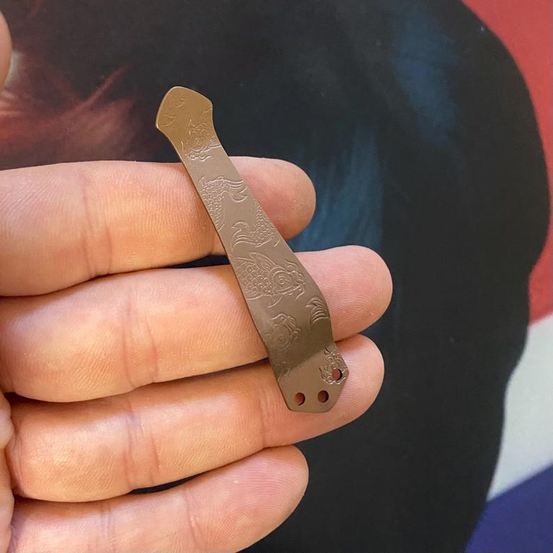 May include: A brown metal pocket clip with a decorative design of fish and waves. The clip has three holes at the bottom.