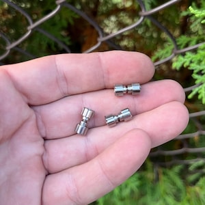 Könnte beinhalten: Drei silberne Metall-Drehwirbel, jeder mit einem kleinen, runden Knopf an jedem Ende. Die Drehwirbel liegen auf der Hand einer Person.