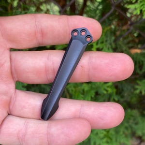May include: A black metal pocket clip with three circular holes at the top and a pointed bottom. The clip is held in a hand against a blurred green background.