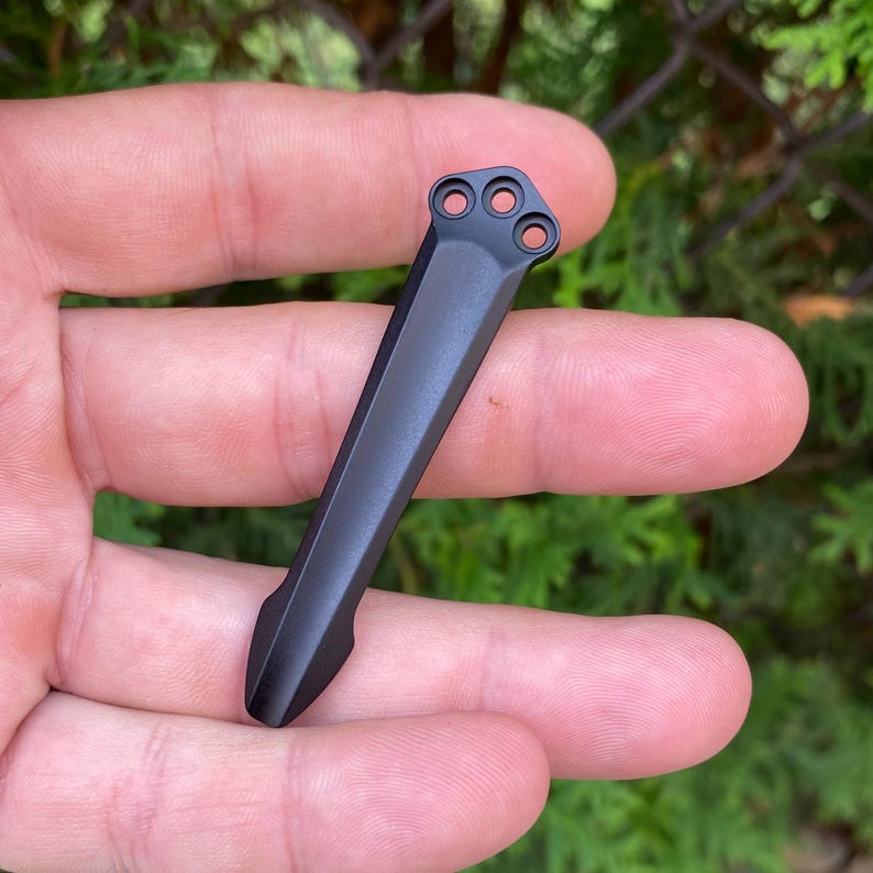 May include: A black metal pocket clip with three circular holes at the top and a pointed bottom. The clip is held in a hand against a blurred green background.