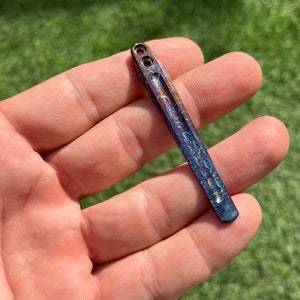 May include: A small, iridescent blue and copper colored metal tool with two holes at the top. The tool has a rounded tip and a textured surface.