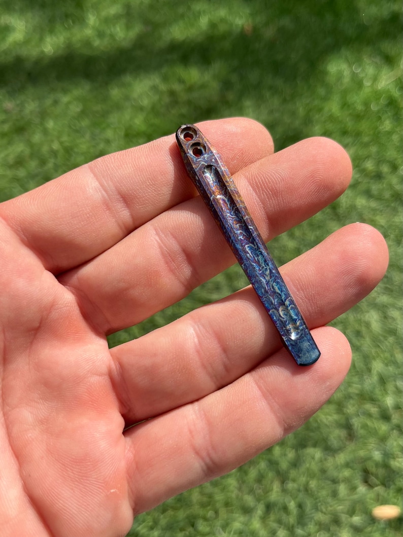 May include: A small, iridescent blue and copper colored metal tool with two holes at the top. The tool has a rounded tip and a textured surface.