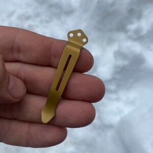 May include: A gold metal pocket clip with a rounded tip and three holes at the top.