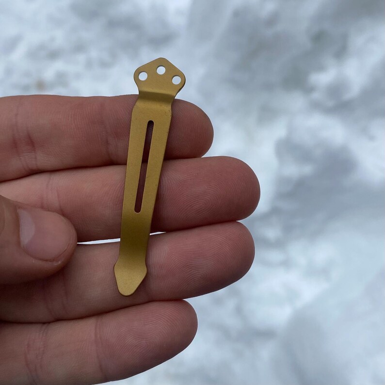 May include: A gold-colored metal pocket clip with three holes at the top and a slot in the middle.