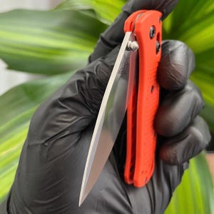 May include: A bright orange folding knife with a silver blade. The knife is held in a black gloved hand, with a green leafy background. The blade is partially open, revealing the sharp edge. The knife has a textured handle.