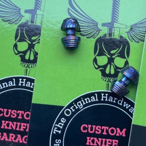 Può includere: Due placche per coltelli in metallo blu e viola con una finitura testurizzata. Le placche sono su uno sfondo verde con un design a teschio e ali. Il testo "The Original Hardware" e "Custom Knife Garage" è visibile.