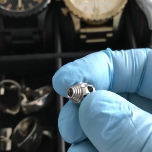 Może przedstawiać: Zbliżenie na srebrny metalowy łącznik do paska zegarka. Łącznik ma gwintowany koniec i mały otwór w środku.