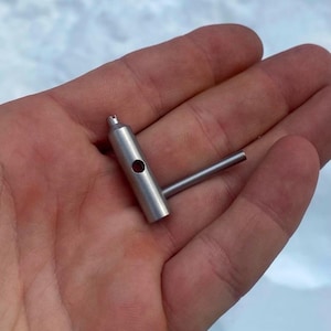 Puede incluir: Una herramienta de metal plateado con un mango en forma de T y un eje largo y delgado. La herramienta tiene un pequeño agujero en el mango y una punta puntiaguda.