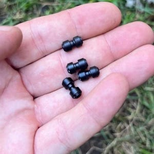 May include: Four small, black, dumbbell-shaped connectors are displayed in a person's open palm. The connectors have a cylindrical shape with a slightly wider center section and are made of a matte black material. The background is blurred, suggesting an outdoor setting.