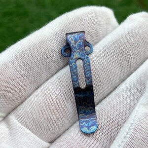 May include: A blue titanium pocket clip with a swirling pattern. The clip has two holes on the top and a curved shape.