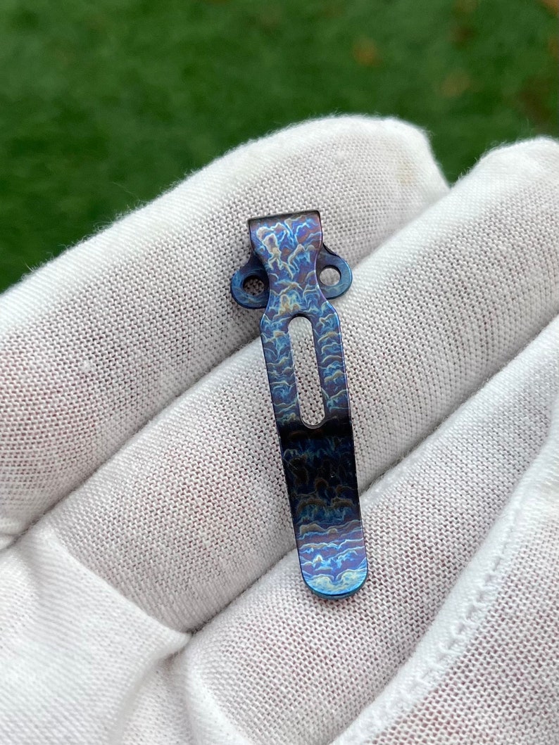 May include: A blue titanium pocket clip with a swirling pattern. The clip has two holes on the top and a curved shape.