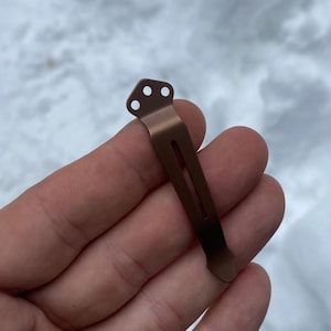 May include: A bronze-colored pocket clip with three small holes at the top and a long, narrow slot. The clip is designed to attach to a pocket or other item. The clip is made of metal and has a smooth finish.