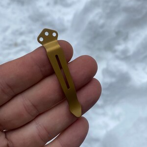 May include: A gold-colored metal pocket clip with three holes at the top and a slot in the middle.