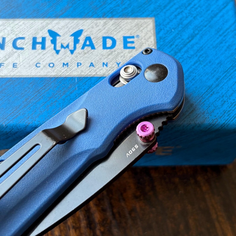 Puede incluir: Una navaja azul con una hoja negra y un clip de bolsillo negro. La navaja tiene un tornillo de acento rosa y el texto "S90V" en la hoja. La navaja est&aacute; colocada sobre una caja azul con la marca "Benchmade".