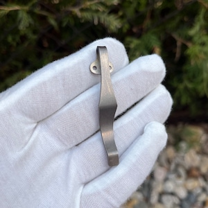May include: A silver-toned metal pocket clip with a curved design. The clip has two small holes at the top.