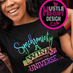 May include: Two women are smiling and making a heart shape with their hands. The woman in the front is wearing a black crop top with a rainbow design that says "Synchronicity a Wink from the Universe Hustle Heart Design Co."