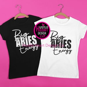 Puede incluir: Dos camisetas, una negra y otra blanca, con el texto "Big Aries Energy" impreso en ellas. El texto está en una fuente blanca y negra en la camiseta negra y en una fuente negra en la camiseta blanca. Las camisetas están colgadas en perchas de madera sobre un fondo rosa.