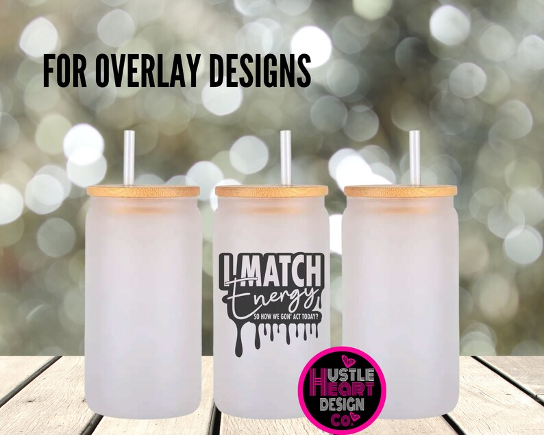 May include: Three frosted glass tumblers with bamboo lids and clear straws. The tumbler on the right has a black graphic that says "I Match Energy" with dripping black paint. The graphic also says "So How We Gon Act Today?" in smaller text. The bottom of the tumbler has a pink heart with the text "Hustle Heart Design Co."