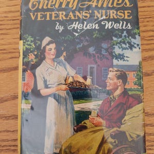 May include: Vintage book cover for "Cherry Ames, Veterans' Nurse" by Helen Wells. The cover features a nurse in a white uniform serving food to a man in a wheelchair. The title is in gold lettering.
