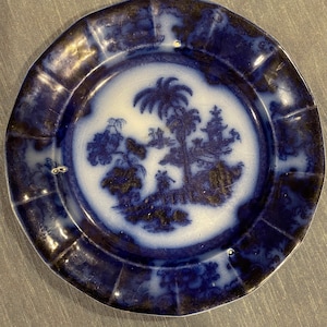 May include: A small, round, antique plate with a blue and white design. The center features a detailed landscape scene with trees and figures. The rim has a scalloped edge and a darker blue hue.
