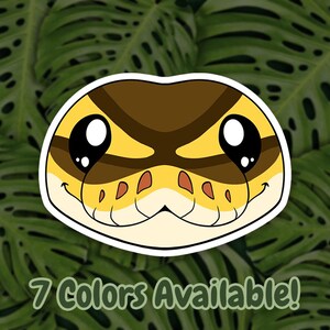 May include: A sticker featuring a cartoon snake head with large, expressive eyes. The snake has a yellow and brown pattern with a white outline. Text at the bottom reads "7 Colors Available!" The background is a pattern of green leaves.