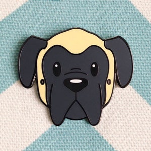 May include: A black and yellow enamel pin featuring a cartoon illustration of a dog's head. The dog has a short, yellow, fluffy top and a black face with a black nose and large, round eyes.