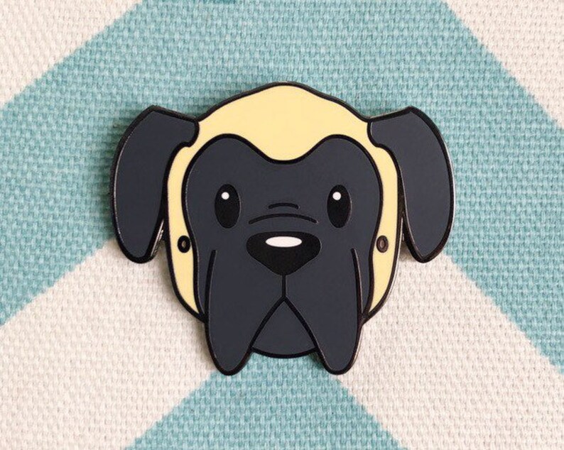 May include: A black and yellow enamel pin featuring a cartoon illustration of a dog's head. The dog has a short, yellow, fluffy top and a black face with a black nose and large, round eyes.