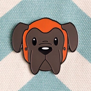 May include: A brown and gray enamel pin of a dog wearing an orange helmet. The dog has a black nose and large, round eyes.