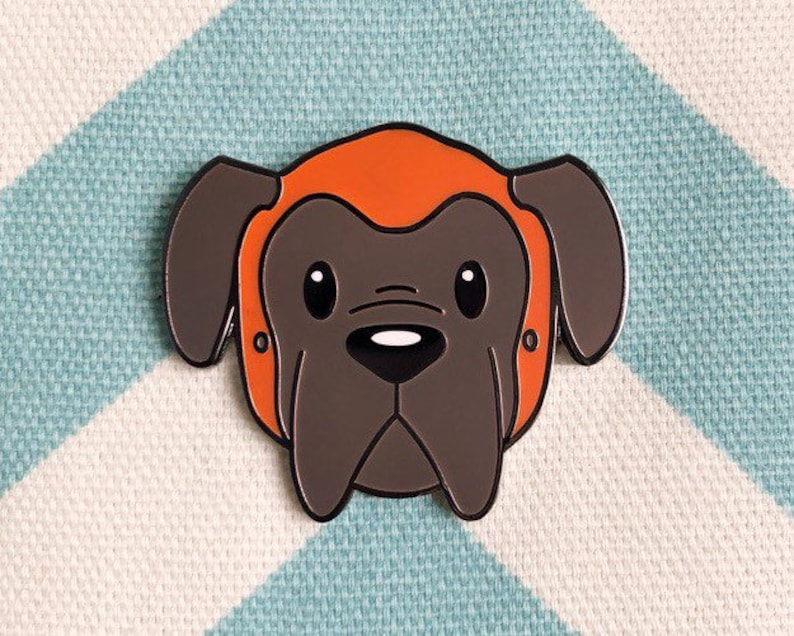 May include: A brown and gray enamel pin of a dog wearing an orange helmet. The dog has a black nose and large, round eyes.