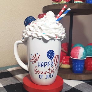 May include: A white mug with a patriotic "Happy Fourth of July" design, topped with whipped cream and festive decorations. It sits on a red stand, with cupcakes and snow cones on a tiered tray in the background.