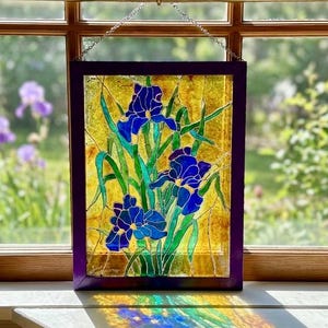 Puede incluir: Un panel rectangular de vidriera que muestra tres flores de iris azules con tallos y hojas verdes sobre un fondo ámbar texturizado. La pieza está enmarcada en color morado oscuro y cuelga de una cadena plateada frente a una ventana.