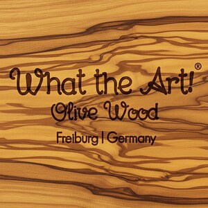 Op de afbeelding: Close-up van een houten oppervlak met de tekst "What the Art!" en "Olive Wood" in donkerbruin. De tekst "Freiburg | Germany" is ook zichtbaar. Het houtnerfpatroon is een mix van licht- en donkerbruin.