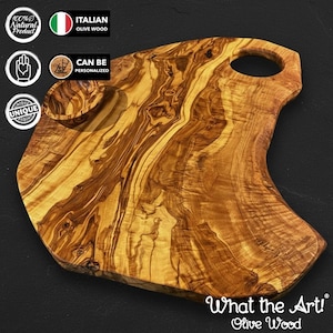 Puede incluir: Tabla de cortar de madera de olivo con un patrón de vetas naturales. La tabla tiene un cuenco pequeño y un asa con un agujero. El texto "What the Art! Olive Wood" es visible. Hecho de madera de olivo italiana.