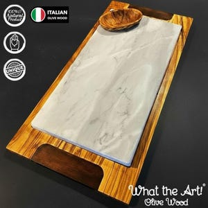 Peut inclure: Un plateau de service rectangulaire avec un centre blanc effet marbre et un cadre en bois. Un petit bol en bois est posé sur le plateau. Le texte "What the Art! Olive Wood" est visible.