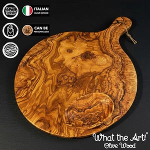 May include: A large, round olive wood cutting board with a handle and a smaller, attached dish. The board has a natural wood grain pattern and is labeled "What the Art! Olive Wood." The image also includes text indicating it is Italian olive wood and a unique, natural product.