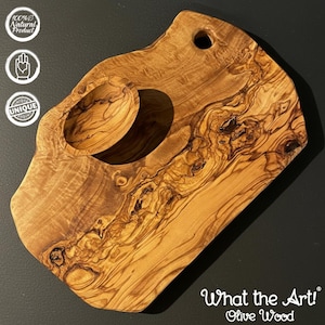 Op de afbeelding: Olijfhouten snijplank met een uniek ontwerp. Het heeft een ingebouwde kom en een gat om op te hangen. De houtnerf is zichtbaar, met natuurlijke tinten. De tekst "What the Art! Olive Wood" is aanwezig.