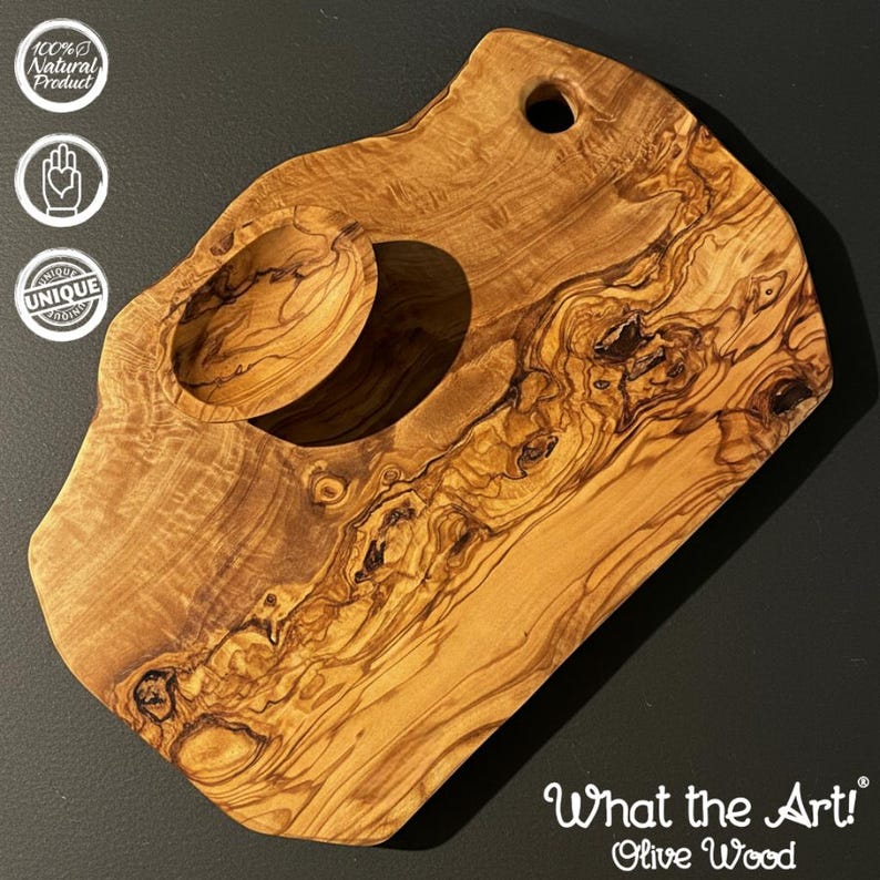 Op de afbeelding: Olijfhouten snijplank met een uniek ontwerp. Het heeft een ingebouwde kom en een gat om op te hangen. De houtnerf is zichtbaar, met natuurlijke tinten. De tekst "What the Art! Olive Wood" is aanwezig.