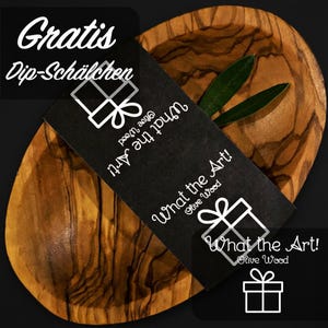 Op de afbeelding: Een houten olijvenschaal met een zwarte kaart met de tekst "What the Art! Olive Wood" in witte letters. De kom heeft een natuurlijk houtnerfpatroon. De kaart is over de kom geplaatst met groene bladeren.