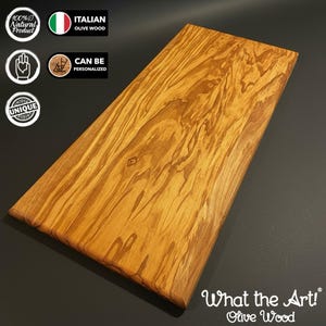 May include: A rectangular olive wood serving board featuring a prominent natural grain pattern with swirling light and dark brown tones. The board has smooth, beveled edges. Text on the image includes 100% Natural Product, Italian Olive Wood, Can Be Personalized, Unique, and What the Art! Olive Wood.