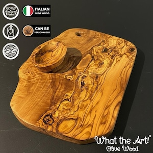 May include: A wooden serving board with a small bowl, crafted from Italian olive wood. The board has a natural, organic shape with unique wood grain patterns. The text "What the Art! Olive Wood" is visible.