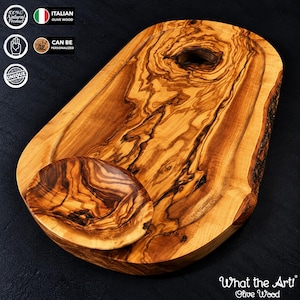 Op de afbeelding: Een ovaal serveerbord van olijfhout met een natuurlijk nerfpatroon, een donkere knoest en een kleine geïntegreerde ronde kom. Het bord heeft aan één kant een natuurlijke rand en de tekst What the Art! Olive Wood staat onderaan.