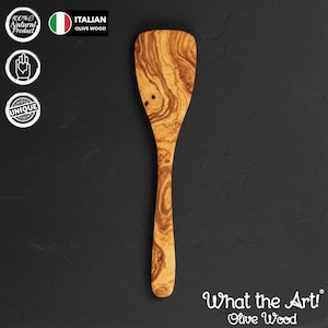 May include: A wooden olive wood spatula with a rounded head and long handle. The spatula has a unique wood grain pattern and is labeled "What the Art! Olive Wood". The image also includes the text "100% Natural Product" and "Italian Olive Wood".