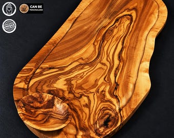 May include: An irregular, oval-shaped olive wood serving board featuring a prominent natural wood grain pattern and a small, integrated circular bowl on one side. The board has a carved perimeter groove and a smooth, polished finish.