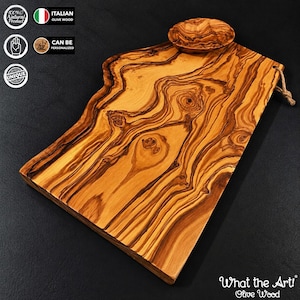 May include: A rectangular olive wood serving board with a natural live edge on one side, featuring a prominent swirling grain pattern in shades of light and dark brown. A small matching wooden bowl sits on the board, and a jute rope loop is attached.