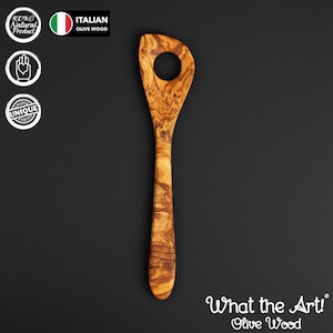 Könnte beinhalten: Ein Kochlöffel aus Olivenholz mit einem Loch im paddelförmigen Kopf. Der Löffel hat ein natürliches Holzmaserungsmuster und einen langen Griff. Der Text "What the Art! Olive Wood" steht unten.