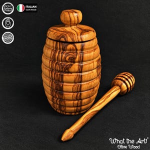 May include: A honey pot crafted from olive wood with a ribbed, barrel-like body and a matching lid with a knob handle. Beside the pot lies a wooden honey dipper with a grooved head. The wood features a distinct, swirling grain pattern in brown.