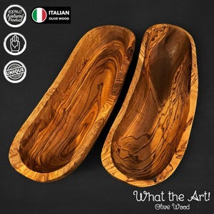 Op de afbeelding: Twee langwerpige houten kommen met een natuurlijk houtnerfpatroon. De kommen zijn gemaakt van Italiaans olijfhout en voorzien van de tekst "What the Art! Olive Wood". De kommen zijn gelabeld als een 100% natuurlijk en uniek product.