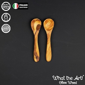 Può includere: Due piccoli cucchiai in legno d'ulivo, di colore marrone chiaro e con venature uniche. Sono presentati su uno sfondo scuro. L'immagine include il testo "What the Art! Olive Wood".
