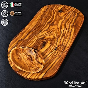 Pode incluir: Uma tábua de servir oval em madeira de oliveira com um padrão de grão natural proeminente e uma pequena taça oval a condizer sobre a sua superfície. A madeira apresenta tons castanhos claros e escuros com nós e texturas orgânicas.
