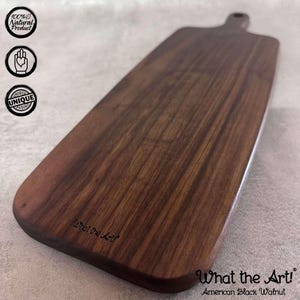 Könnte beinhalten: Ein langes, rechteckiges Servierbrett aus dunkelbraunem amerikanischem Schwarznussbaumholz. Das Brett hat eine abgerundete Kante und einen Griff an einem Ende. Der Text "What the Art!" ist in das Brett graviert.